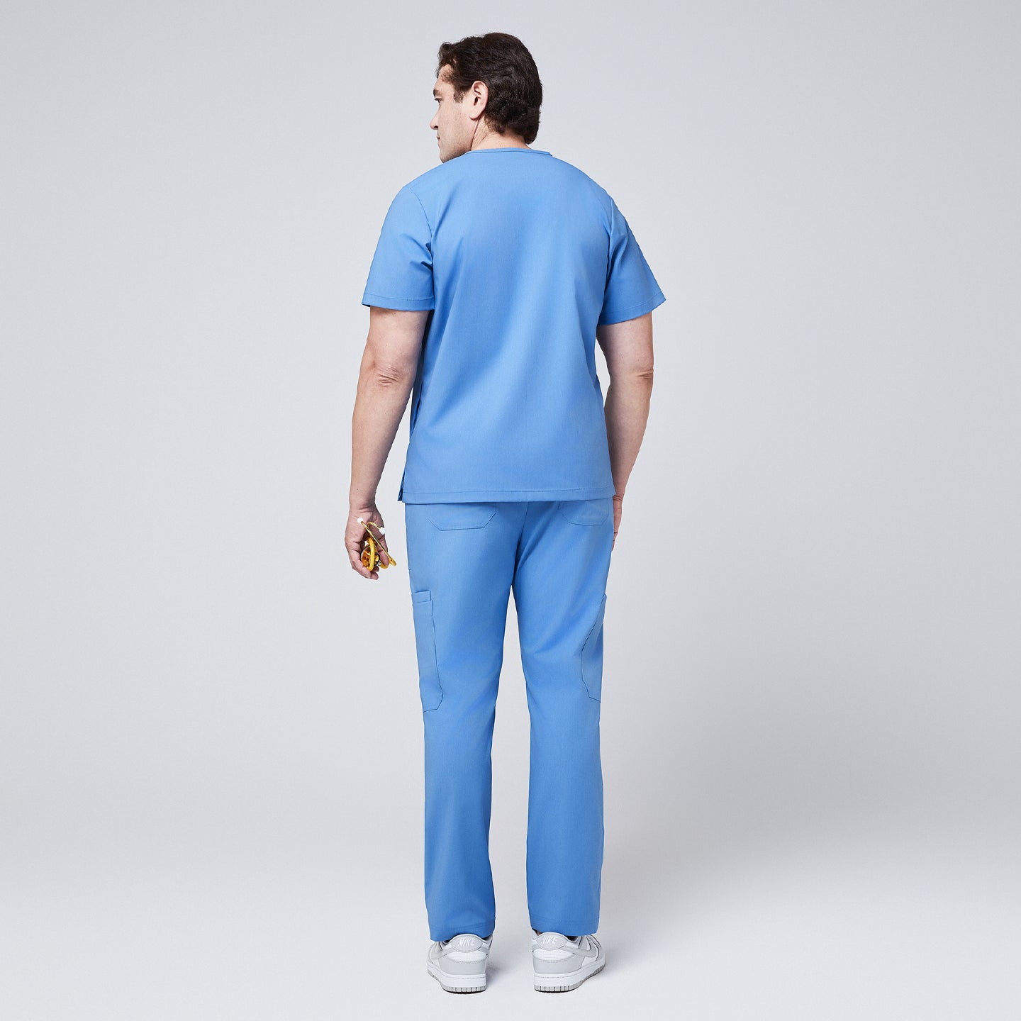 Male model in a ceil blue V-neck scrub set featuring straight-leg pants with functional pockets, holding a yellow stethoscope and wearing white sneakers,Ceil Blue