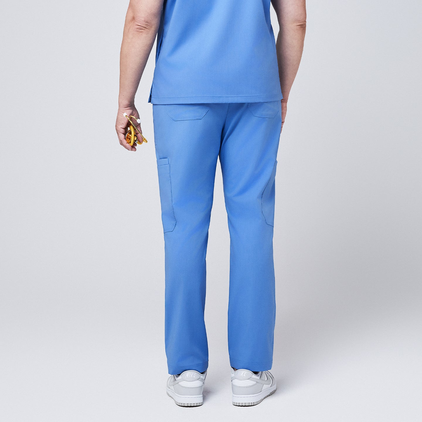 Back view of a person wearing ceil blue straight scrub pants showcasing utility pockets, paired with white sneakers and holding a yellow stethoscope,Ceil Blue