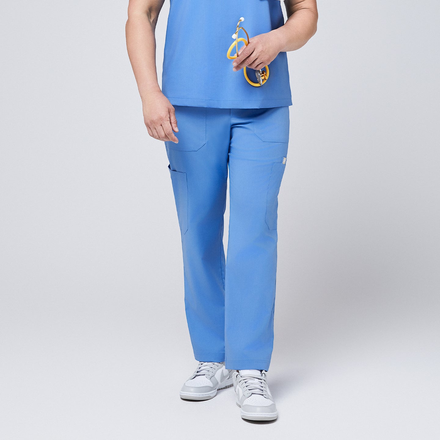 Close-up view of the lower half of a person wearing ceil blue straight scrub pants with multiple utility pockets, paired with white sneakers, holding a yellow stethoscope,Ceil Blue