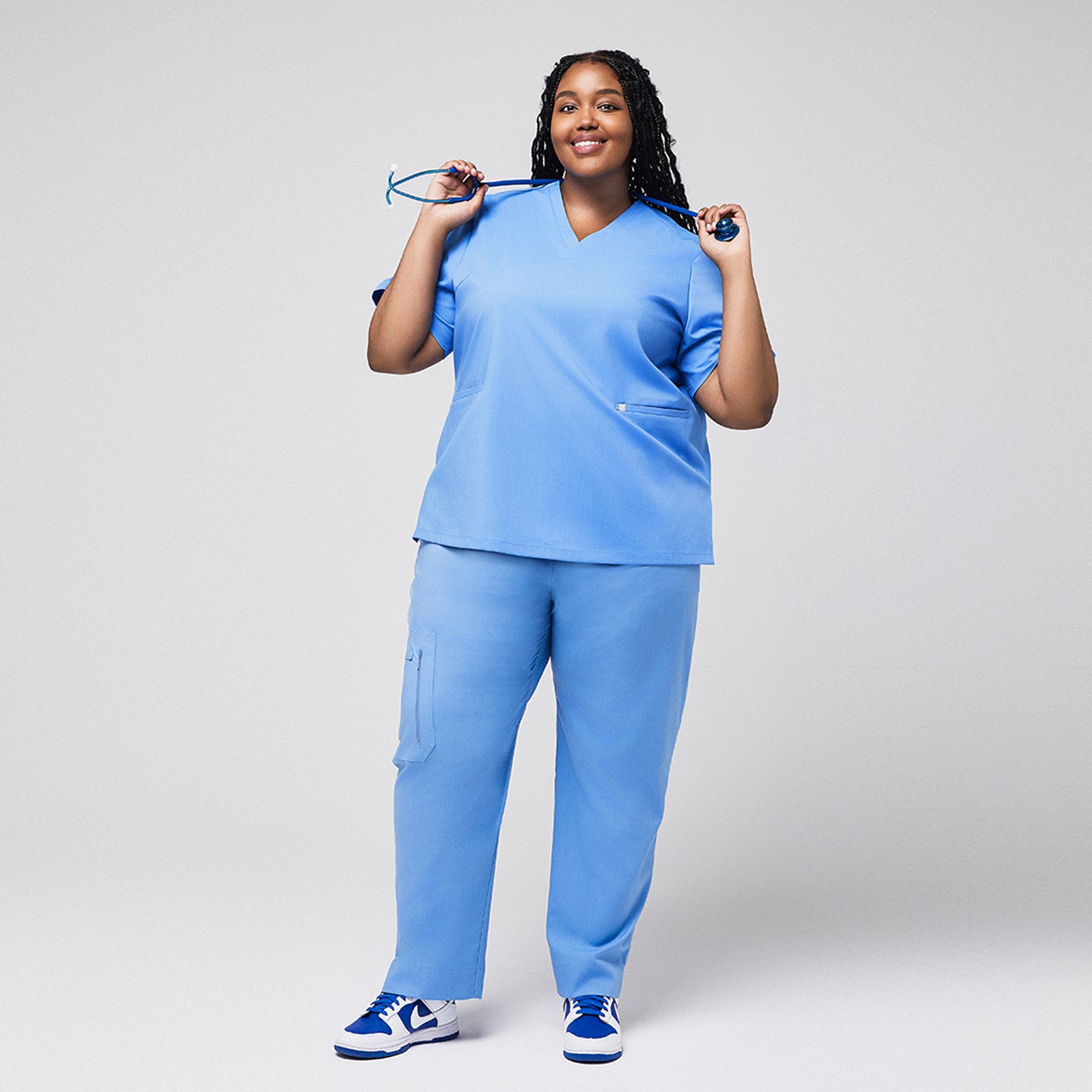 Plus-size healthcare professional wearing a ceil blue scrub set, holding a stethoscope with both hands over the shoulder, smiling with a full-body view,Ceil Blue