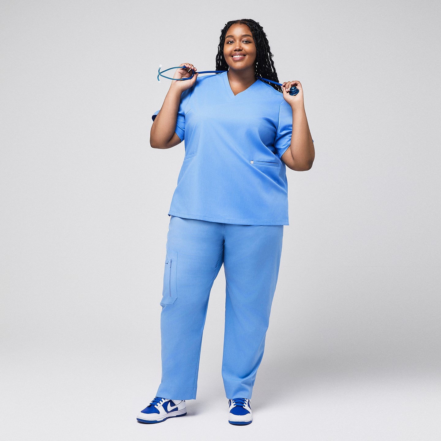 A plus-size model wearing a Ceil Blue Double Pocket Scrub Top and Ceil Blue Zipper Pocket Jogger Scrub Pants, holding a stethoscope behind her neck,Ceil Blue