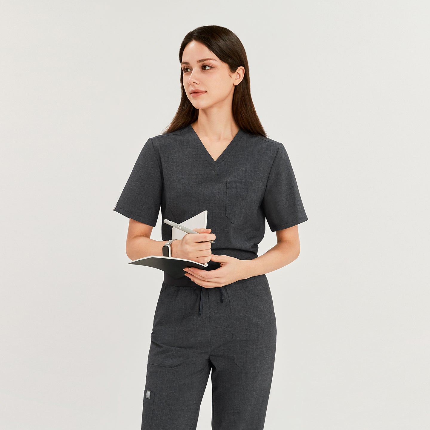 Female model wearing a charcoal gray 3-pocket scrub top, holding a notebook and pen, perfect for healthcare professionals seeking a functional and stylish uniform,Charcoal Gray