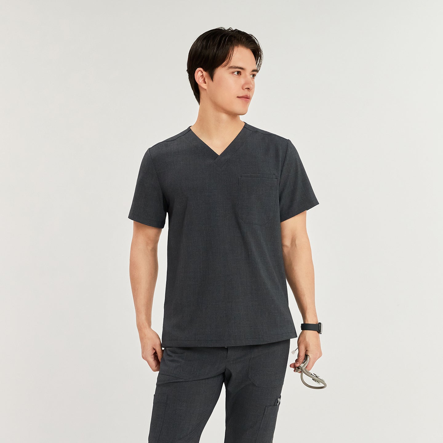 Male model wearing a charcoal gray 3-pocket scrub top, standing and looking to the side. He is holding a stethoscope in his right hand,Charcoal Gray