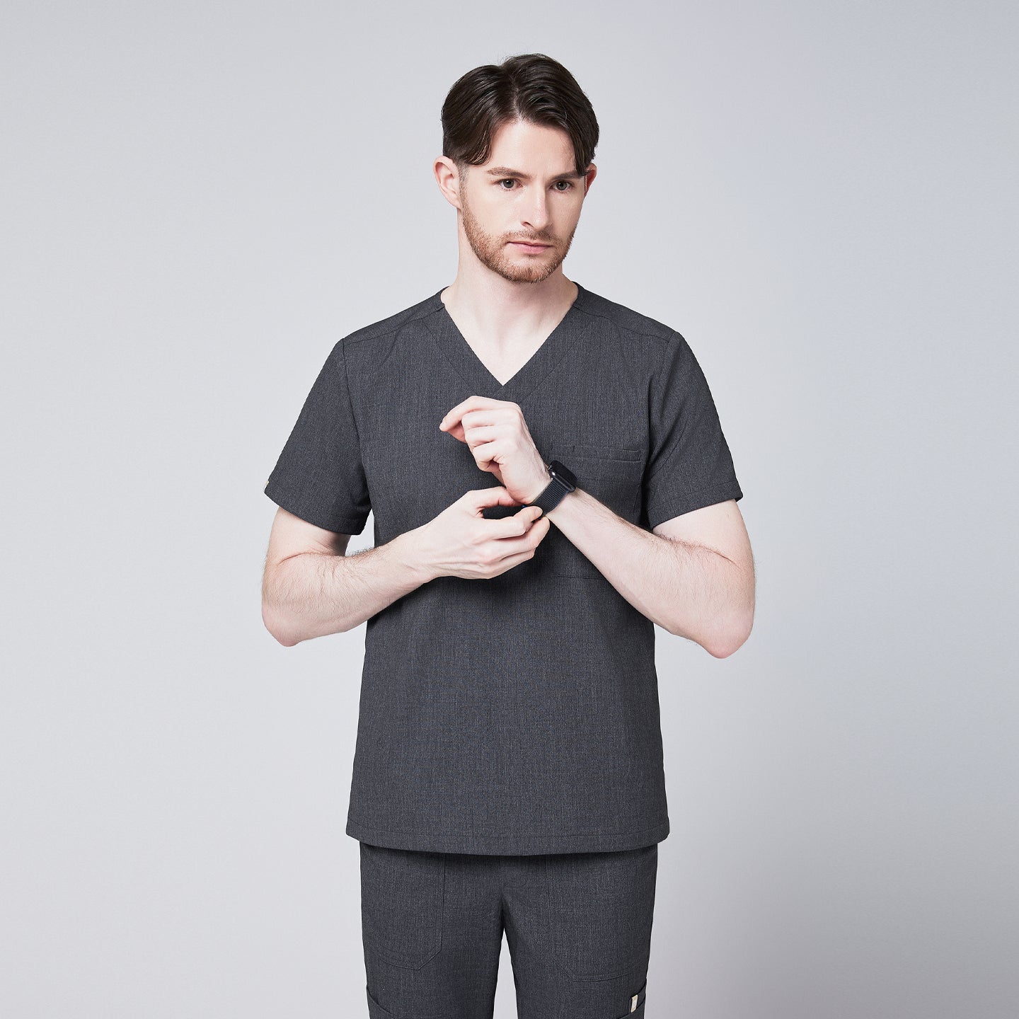 Male healthcare professional wearing charcoal gray V-neck scrub top and pants, adjusting his smartwatch, standing against a plain background,Charcoal-Gray