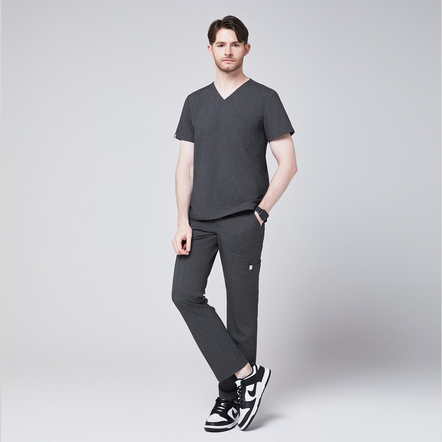 Male healthcare professional wearing a charcoal gray V-neck scrub top and matching jogger pants, with one hand in pocket, standing against a plain background,Charcoal-Gray