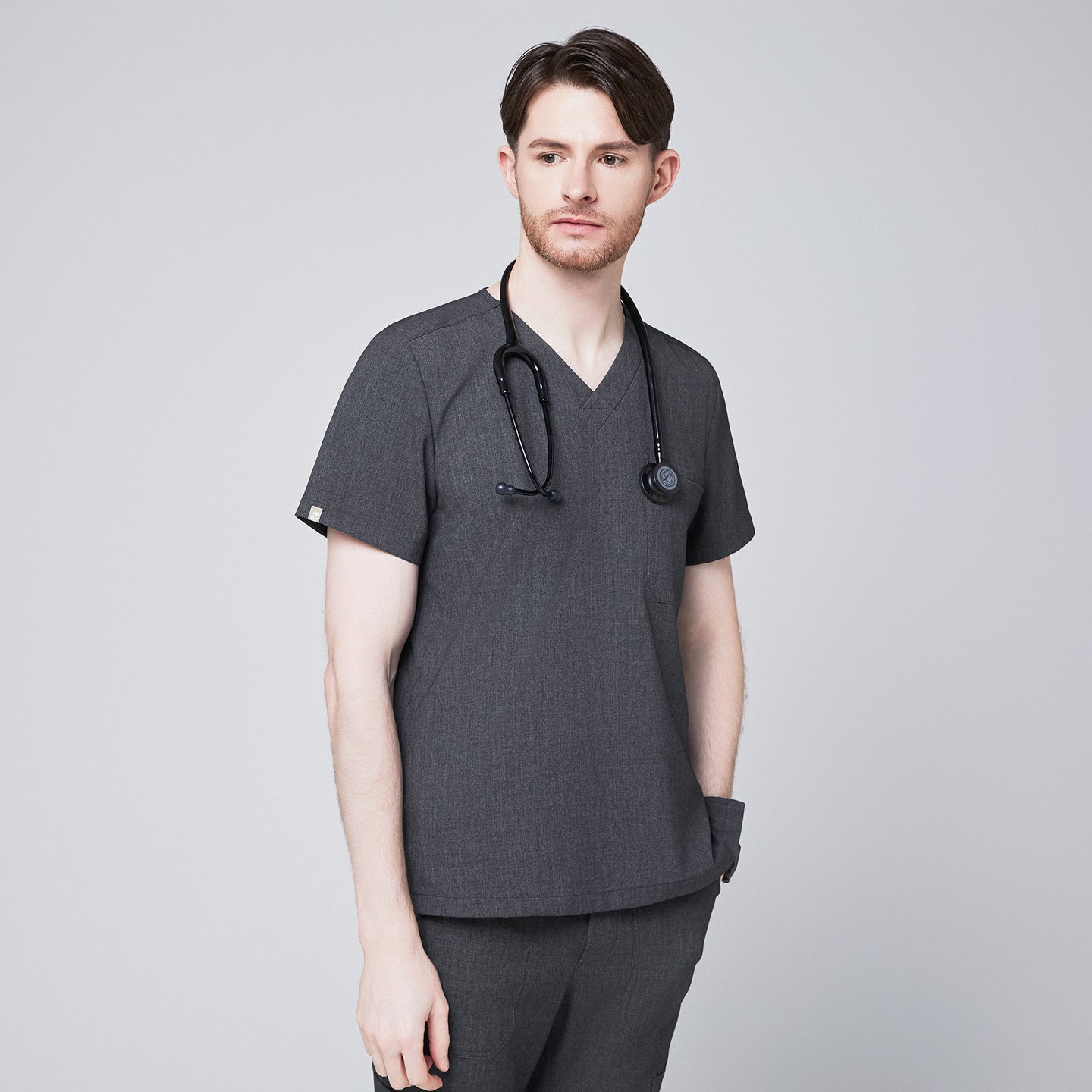 Male healthcare professional in charcoal gray V-neck scrub top and pants, with black stethoscope around neck and hand in pocket, looking to the side,Charcoal-Gray