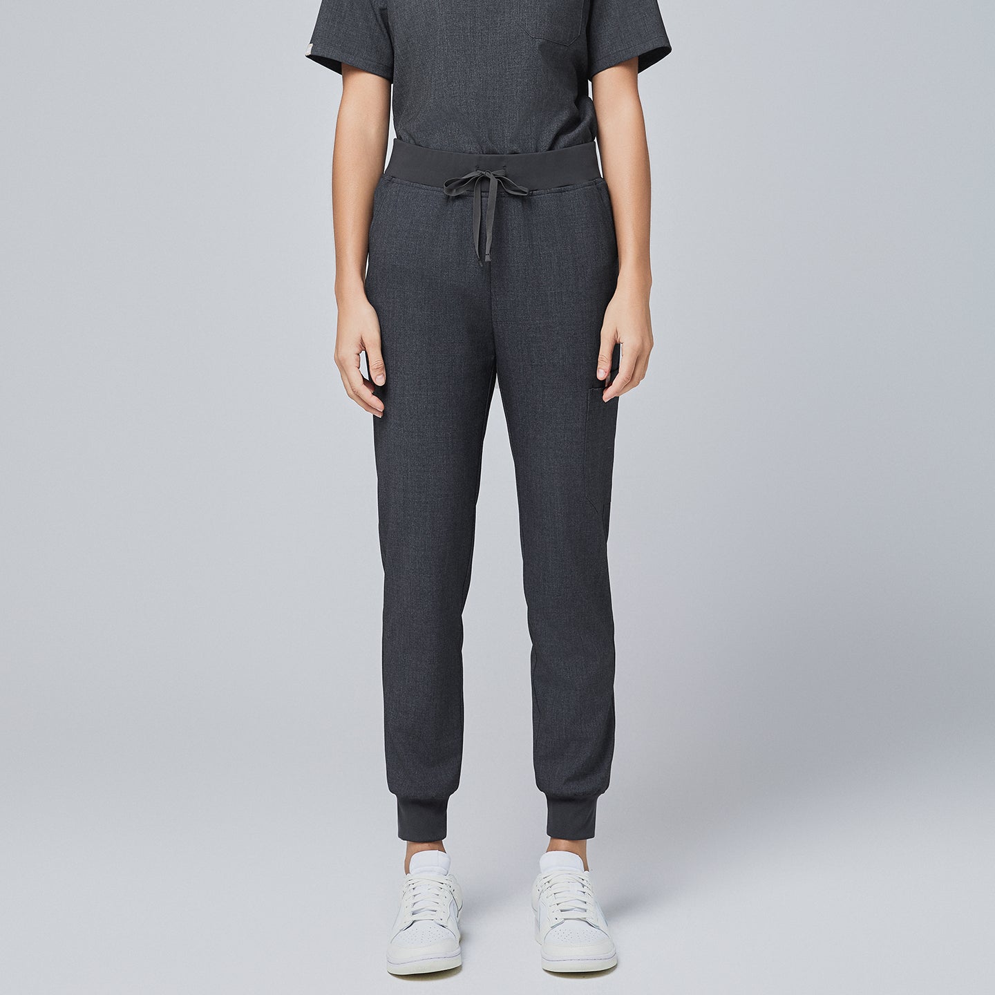 Front view of charcoal gray jogger scrub pants with an elastic waistband, adjustable drawstring, and cuffed ankles, paired with white sneakers,Charcoal Gray