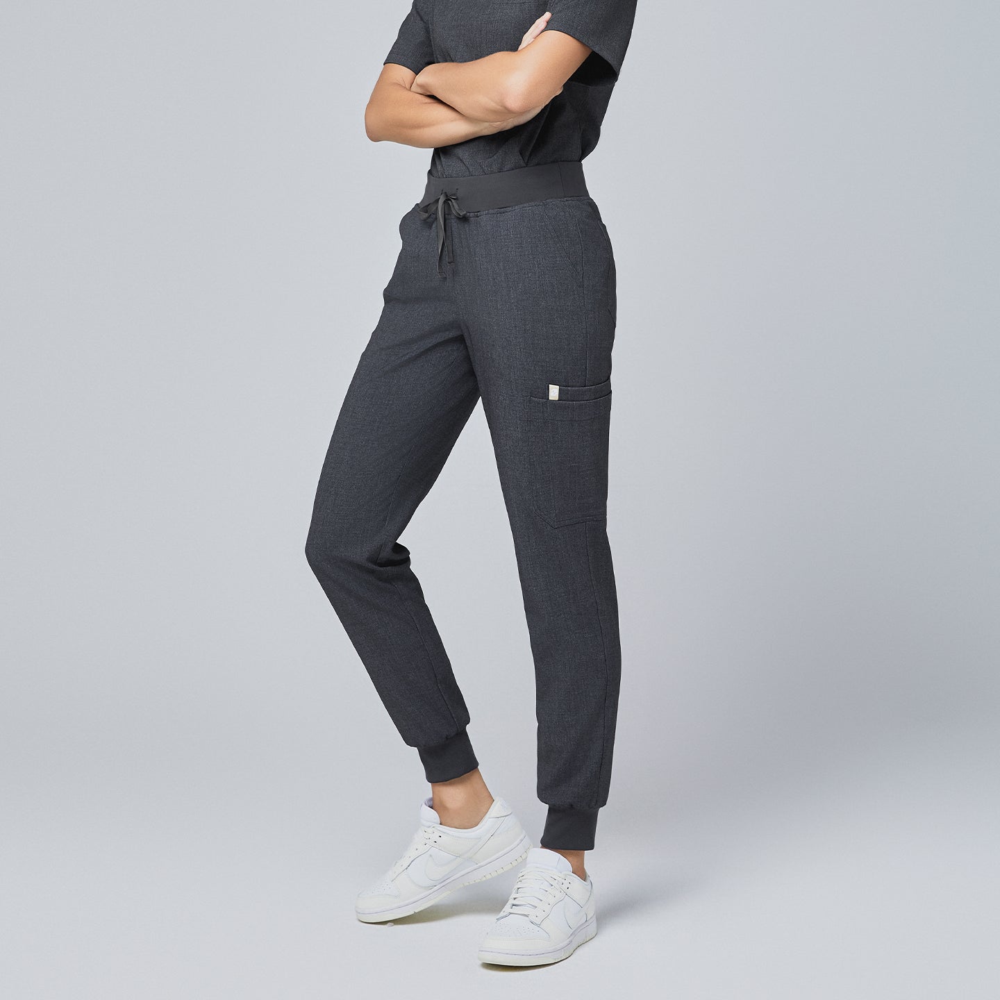 Side view of charcoal gray jogger scrub pants featuring a side pocket, elastic waistband with a drawstring, and cuffed ankles, paired with white sneakers,Charcoal Gray