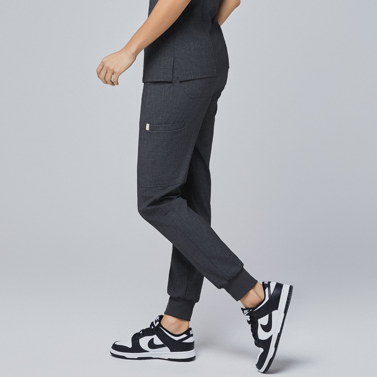 Side angle of charcoal gray jogger scrub pants with a side pocket, elastic cuffs, and a tapered fit, paired with black-and-white sneakers,Charcoal Gray