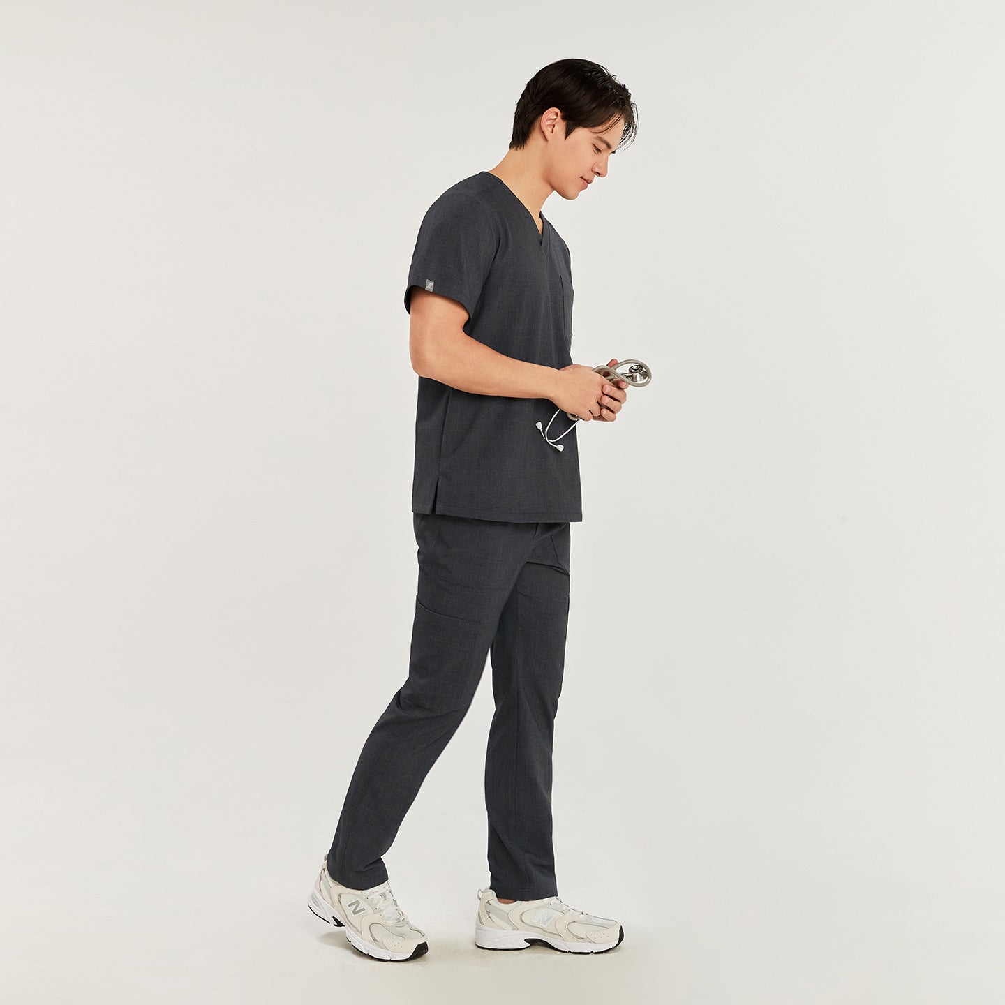 Side view of a model in charcoal gray straight scrub pants with zipper details, matching top, and white sneakers, holding a stethoscope,Charcoal Gray