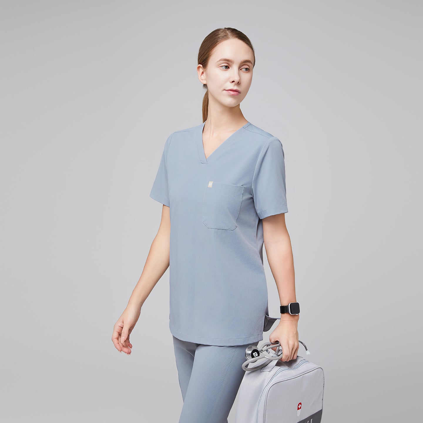 Female healthcare professional in a cool gray scrub set, holding a light gray medical kit bag, walking in a side profile,Cool Gray