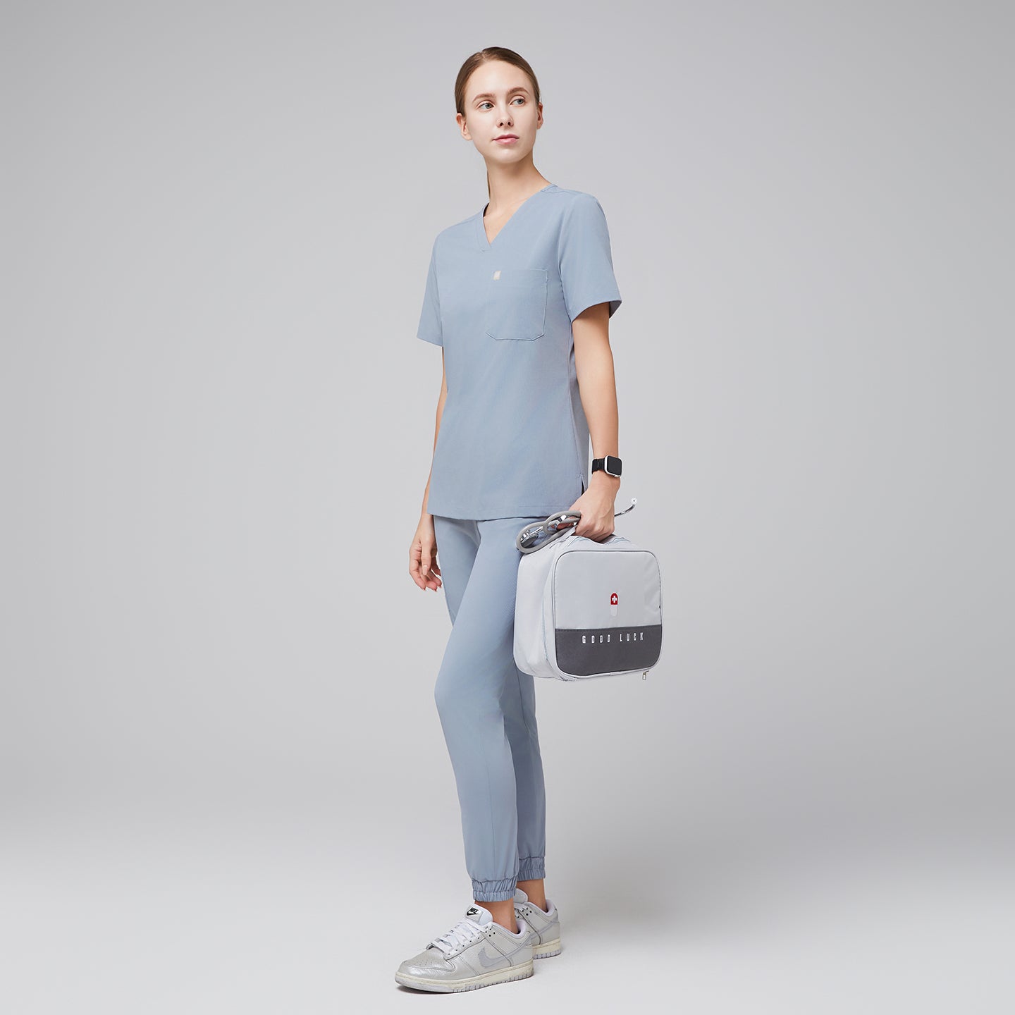 Female healthcare professional in a cool gray scrub set, holding a light gray medical kit bag and wearing silver sneakers, standing in a relaxed pose,Cool Gray