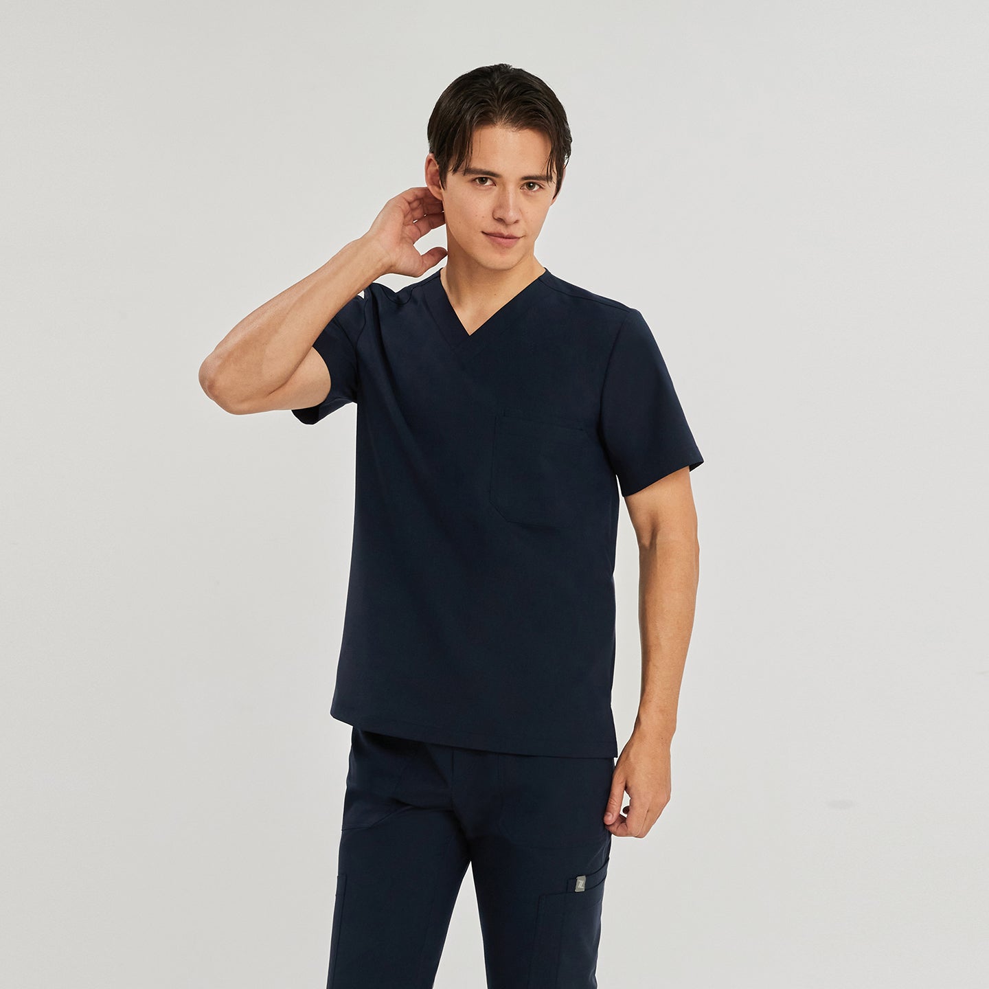 Male model wearing a navy blue scrub top with a V-neck and a single chest pocket, standing against a plain background with his right hand touching his neck,Navy