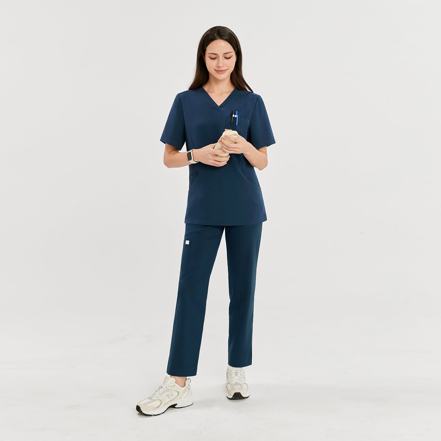 Woman wearing a dark blue soft stretch scrub top and matching pants, holding a hand model, looking down, with pens in the chest pocket and a smartwatch on her wrist,Dark Blue