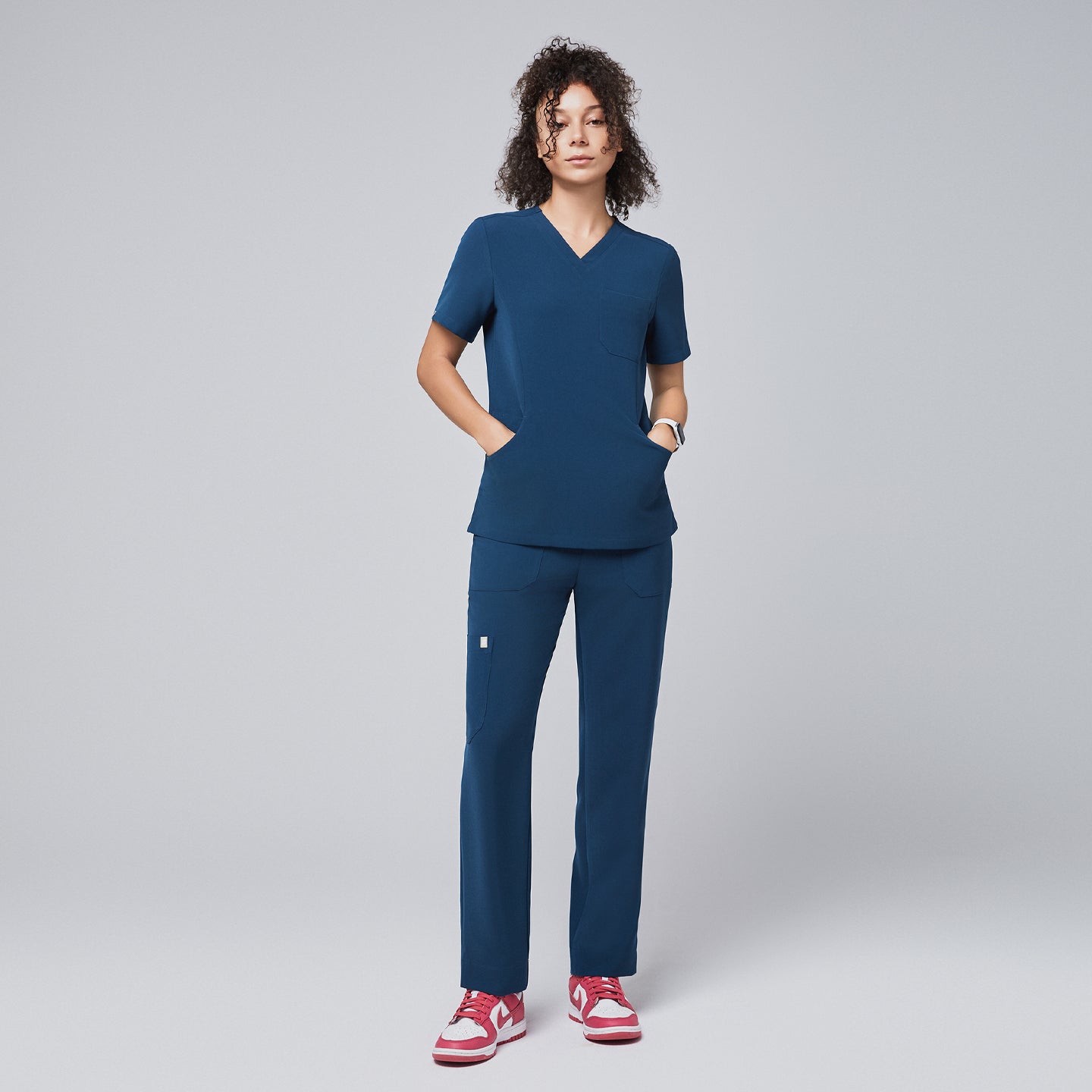 Model dressed in a dark blue V-neck scrub top and matching straight-leg pants with hands in pockets, paired with red and white sneakers, standing against a light gray background,Dark Blue