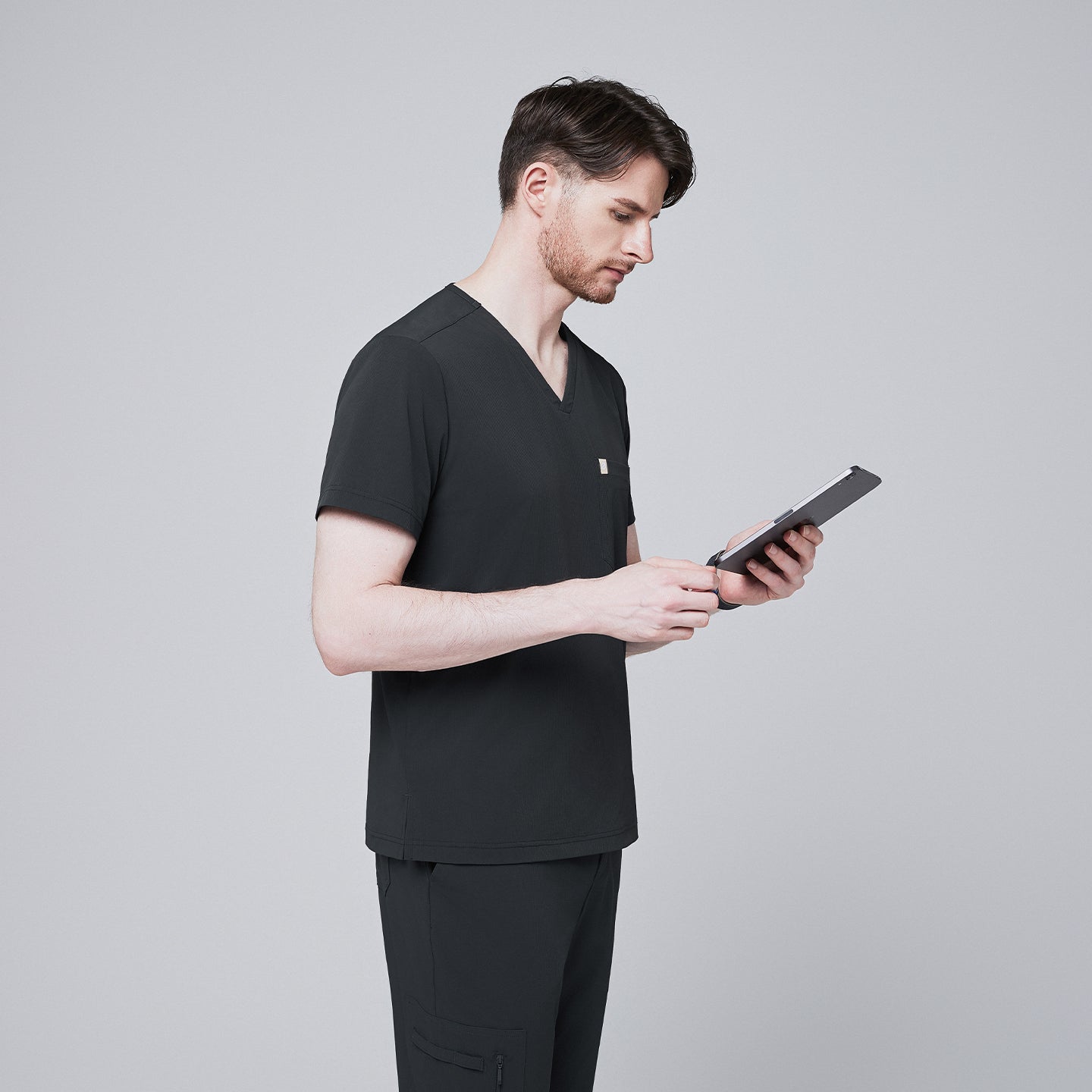 Male model wearing a deep gray V-neck single-pocket scrub top, using a tablet,Deep Gray