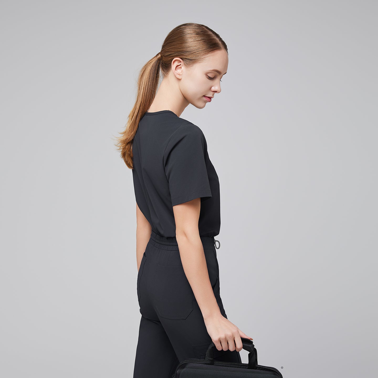 Female healthcare professional in a dark gray scrub set, viewed from the back, holding a small black bag,Deep Gray