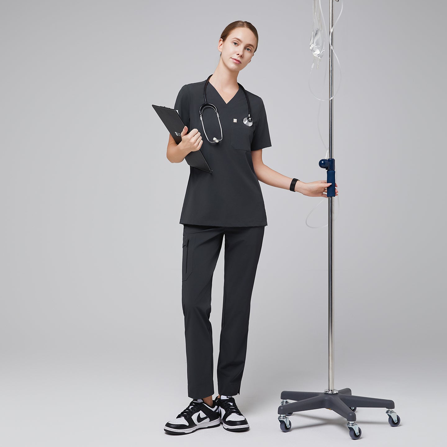 Full-body view of a female healthcare professional wearing a dark gray scrub set, holding a clipboard and IV stand, paired with black sneakers,Deep Gray