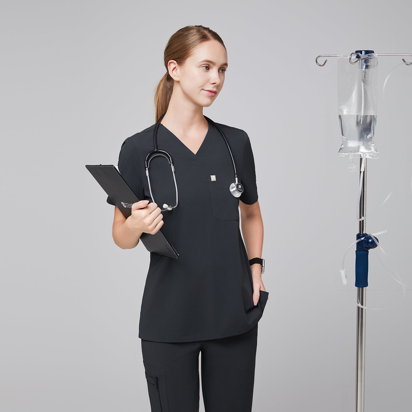 Female healthcare professional wearing a dark gray V-neck scrub top and pants, holding a clipboard with an IV stand in the background,Deep Gray