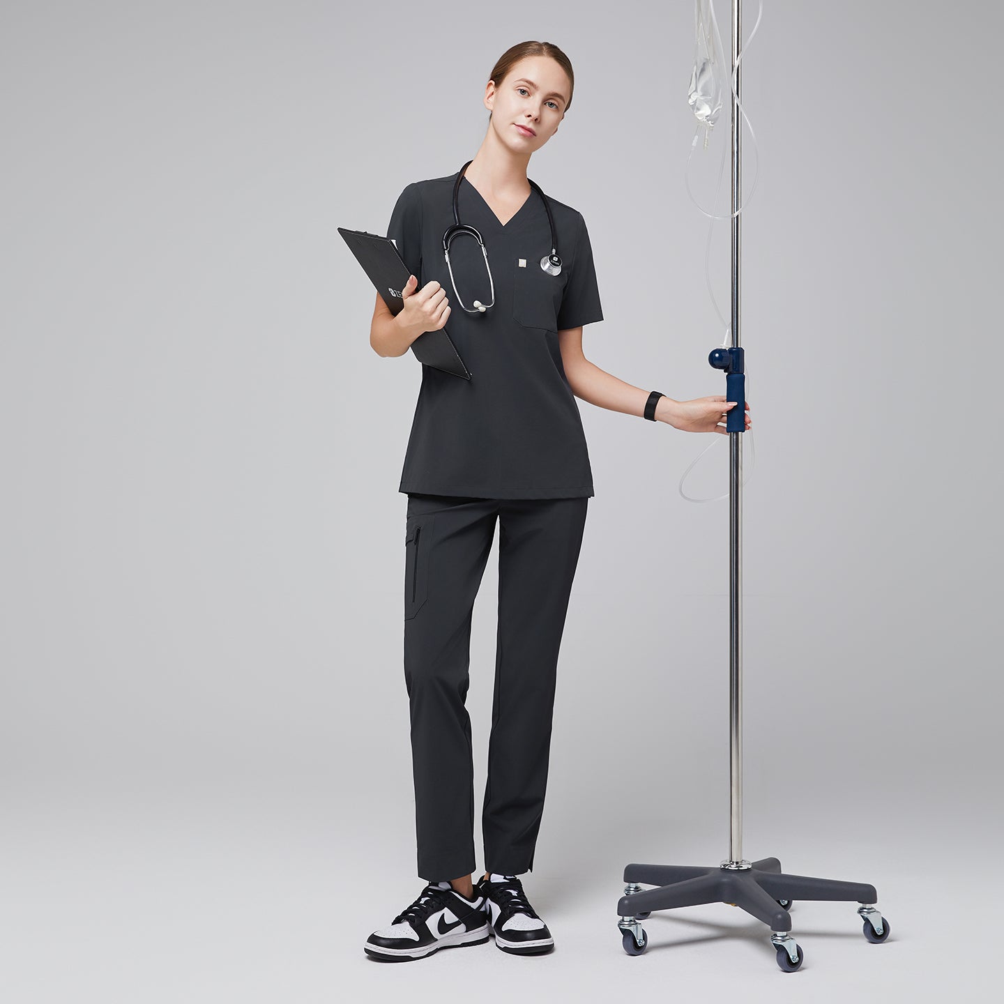 Healthcare professional wearing a scrub set with a V-neck top and zipper pocket pants, holding a clipboard and stethoscope, standing next to an IV pole,Deep Gray