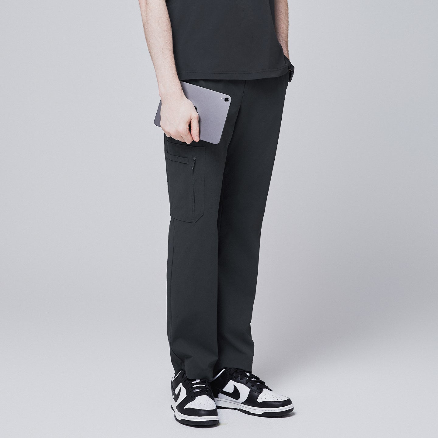 Side view of deep gray straight scrub pants featuring a secure zipper pocket, modeled while holding a tablet, paired with black and white sneakers against a neutral gray background,Deep Gray