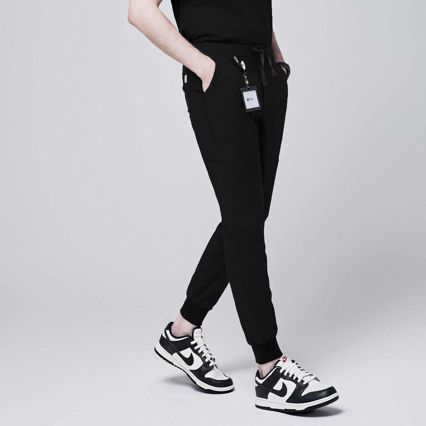 Side angle of black jogger scrub pants with 8 pockets, drawstring waistband, and cuffed ankles, paired with black and white sneakers and a name badge,Eco Black