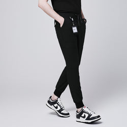 Image of Side angle of black jogger scrub pants with 8 pockets, drawstring waistband, and cuffed ankles, paired with black and white sneakers and a name badge,Eco Black