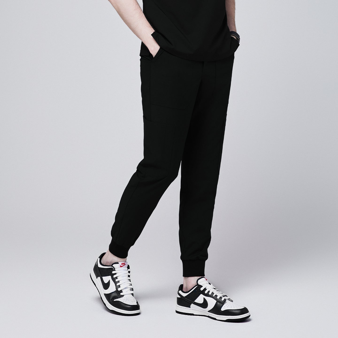 Side view of black jogger scrub pants with 8 pockets, cuffed ankles, and hands in pockets, paired with black and white sneakers,Eco Black