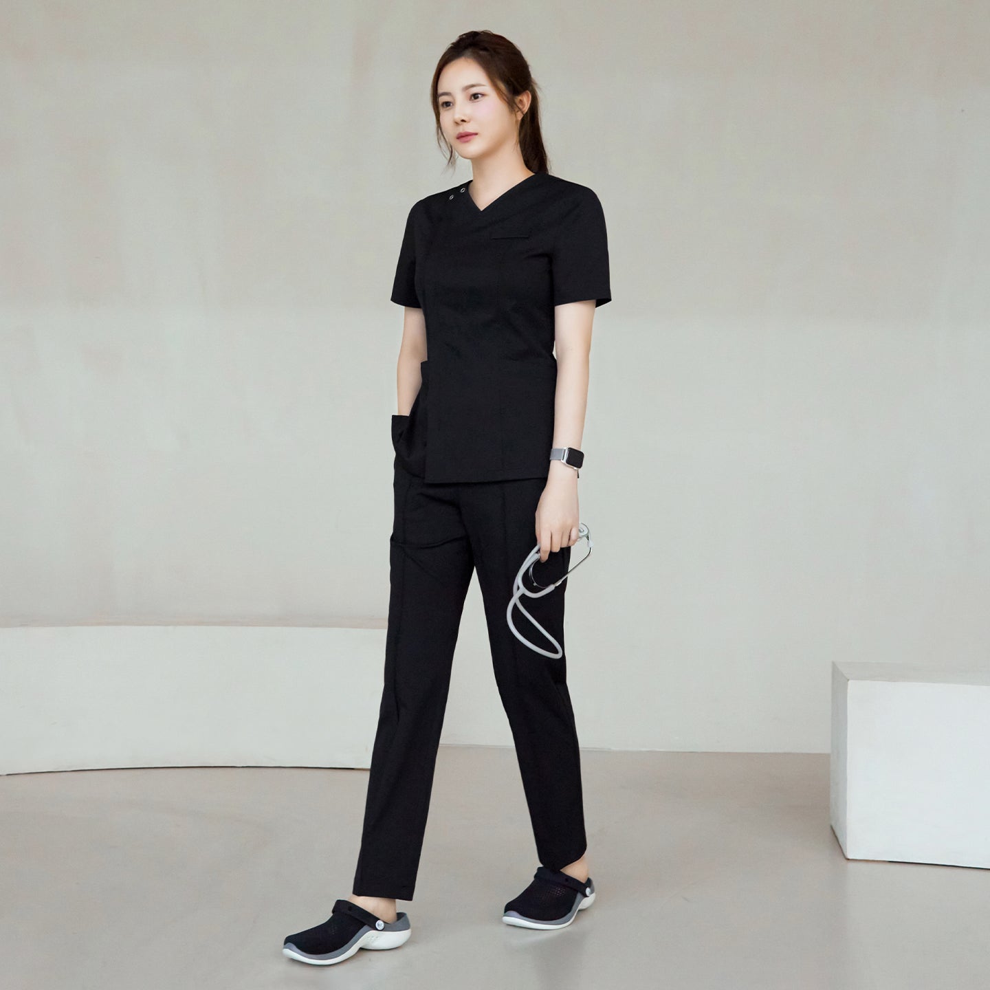 Full body view of a woman wearing Eco Black Line Banding Scrub Pants with a matching top, standing with one hand in her pocket and holding a stethoscope,Eco Black