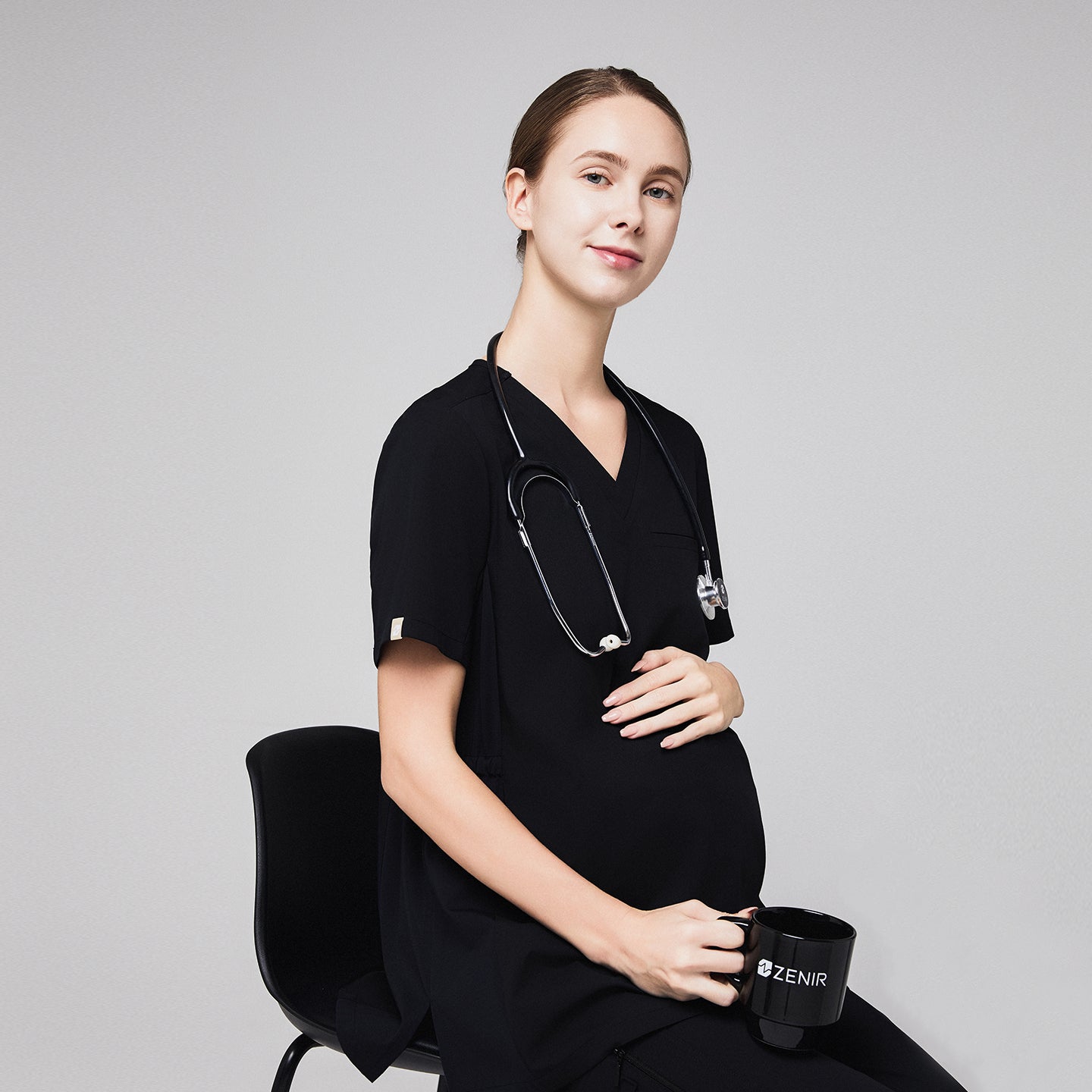 Pregnant healthcare professional in black maternity scrubs, sitting with a stethoscope and holding a black mug,Eco Black