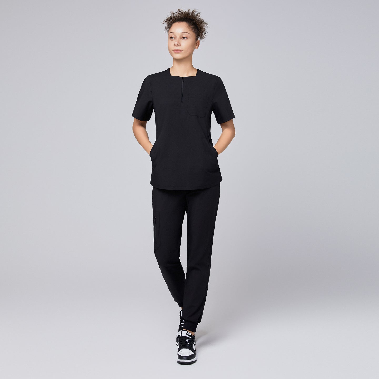 Model wearing a black quarter-zip scrub set with a short-sleeve top featuring a chest pocket and matching jogger-style pants, paired with sneakers for a modern, professional look,Eco Black