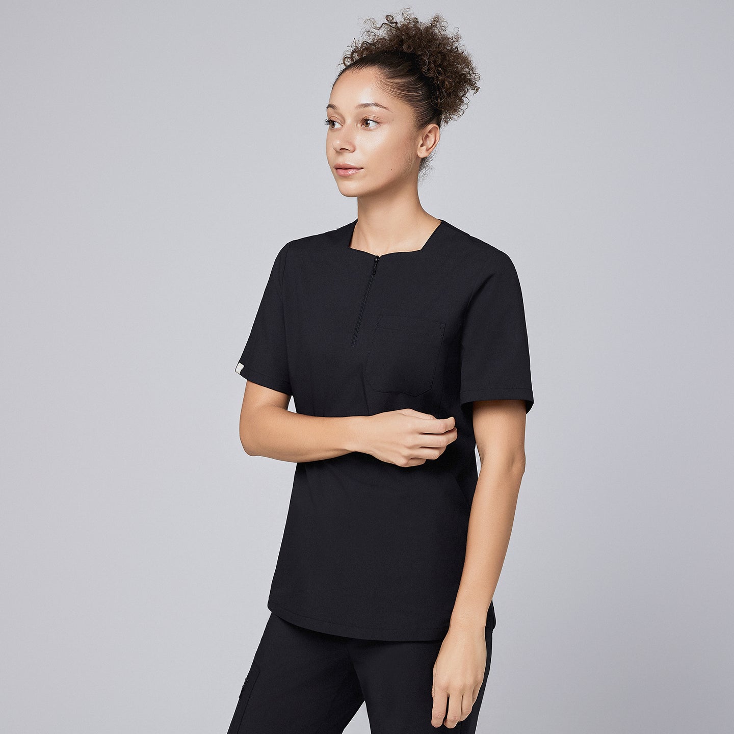 Model wearing eco black scrub top with a quarter zipper, standing with one hand lightly holding the other arm, looking to the side,Eco Black