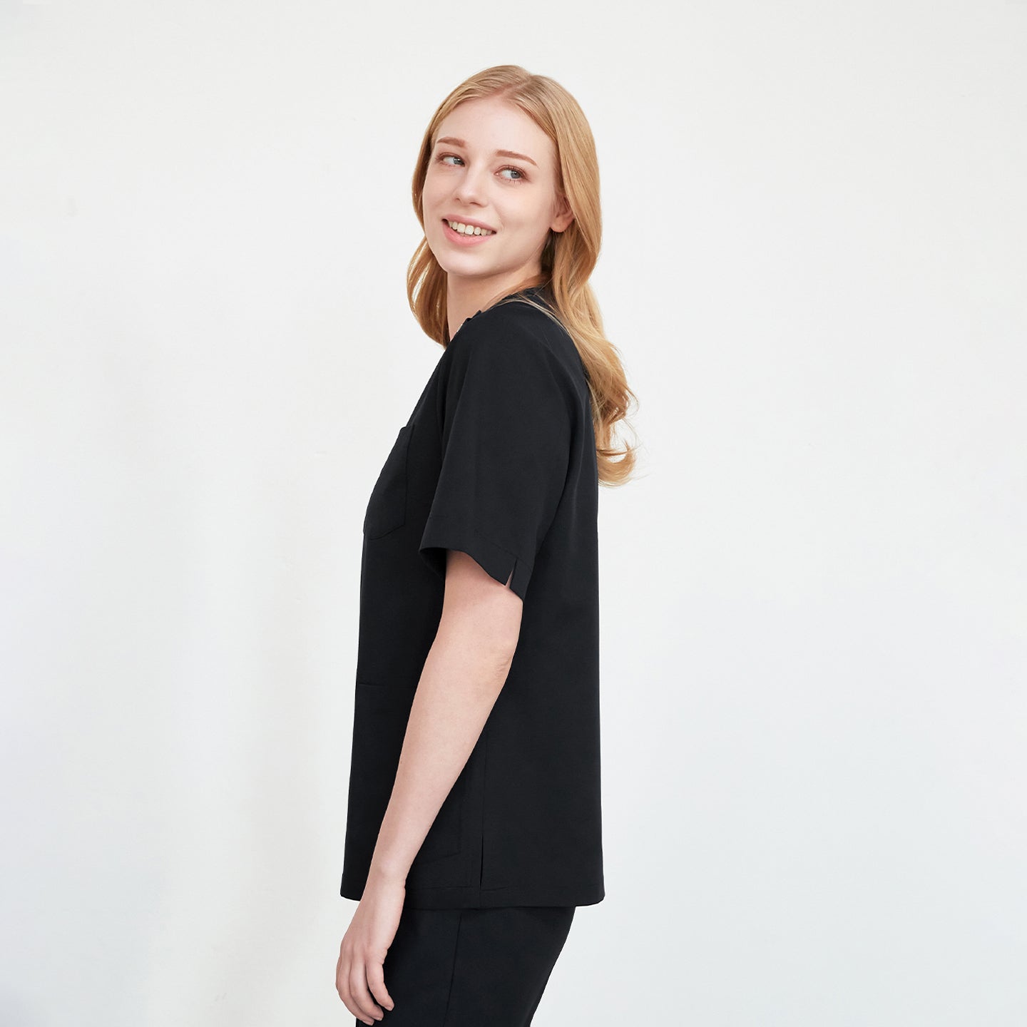 A woman in a black scrub top with buttons on the shoulder, standing and smiling while looking over her shoulder,Eco Black