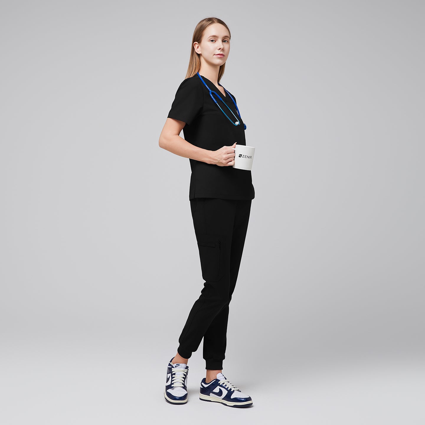 Full-body side view of a model wearing eco black jogger scrub pants with zipper pocket and matching scrub top, holding a Zenir mug and styled with navy sneakers,Eco Black