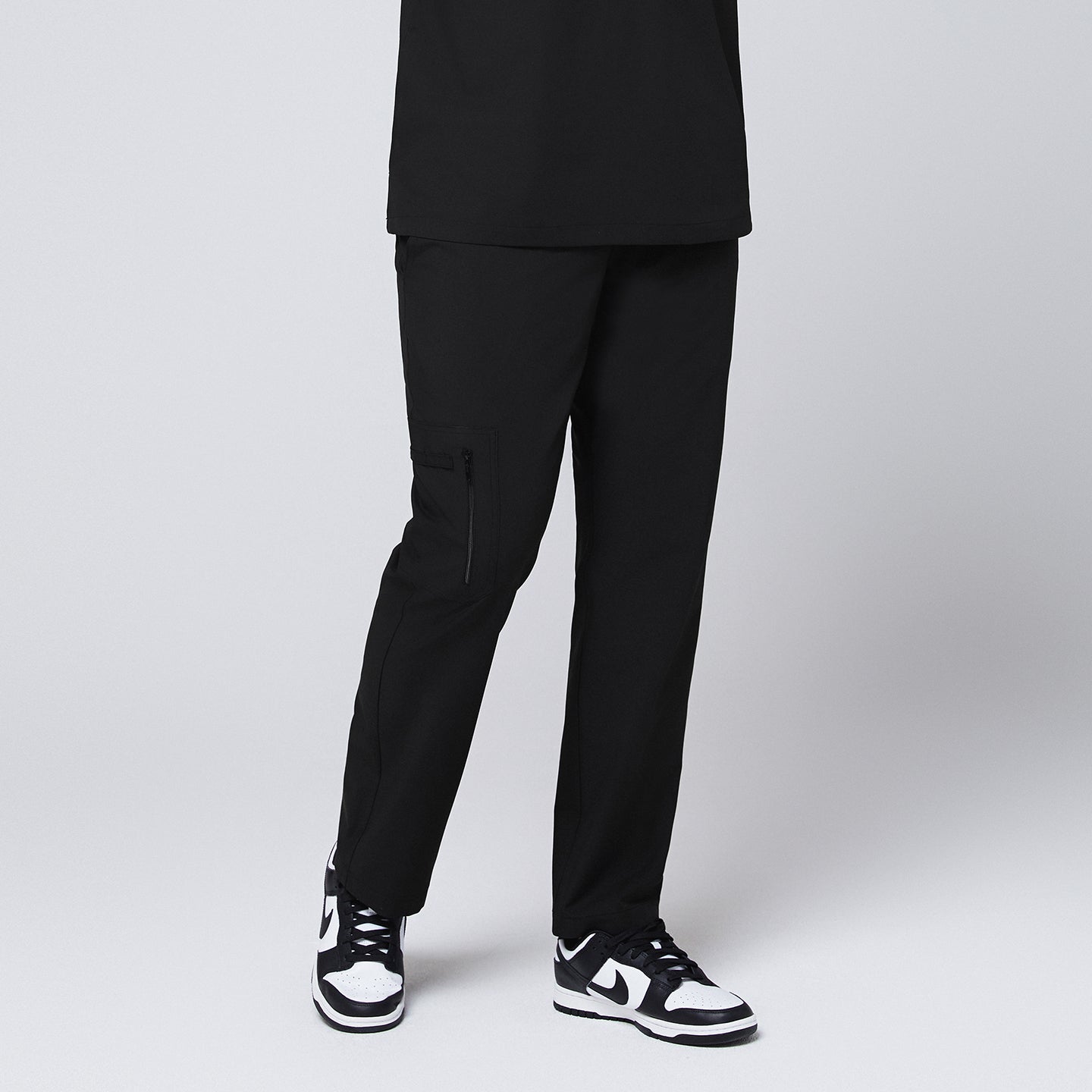 A male model wearing black straight scrub pants with a zipper pocket, paired with black sneakers, standing in a frontal pose,Eco Black