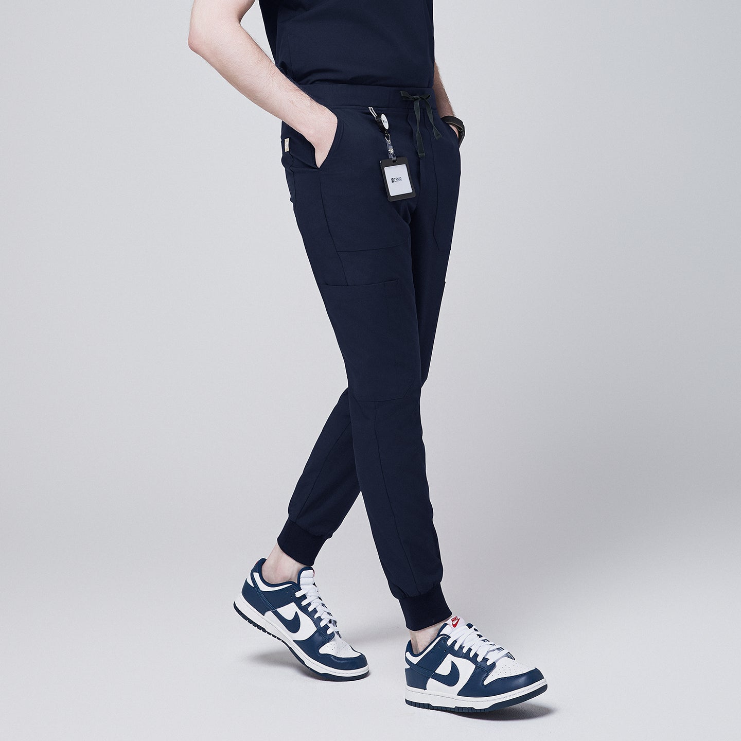 Side angle of navy jogger scrub pants with 8 pockets, drawstring waistband, cuffed ankles, and a name badge, paired with navy sneakers,Eco Navy