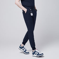 Image of Side angle of navy jogger scrub pants with 8 pockets, drawstring waistband, cuffed ankles, and a name badge, paired with navy sneakers,Eco Navy