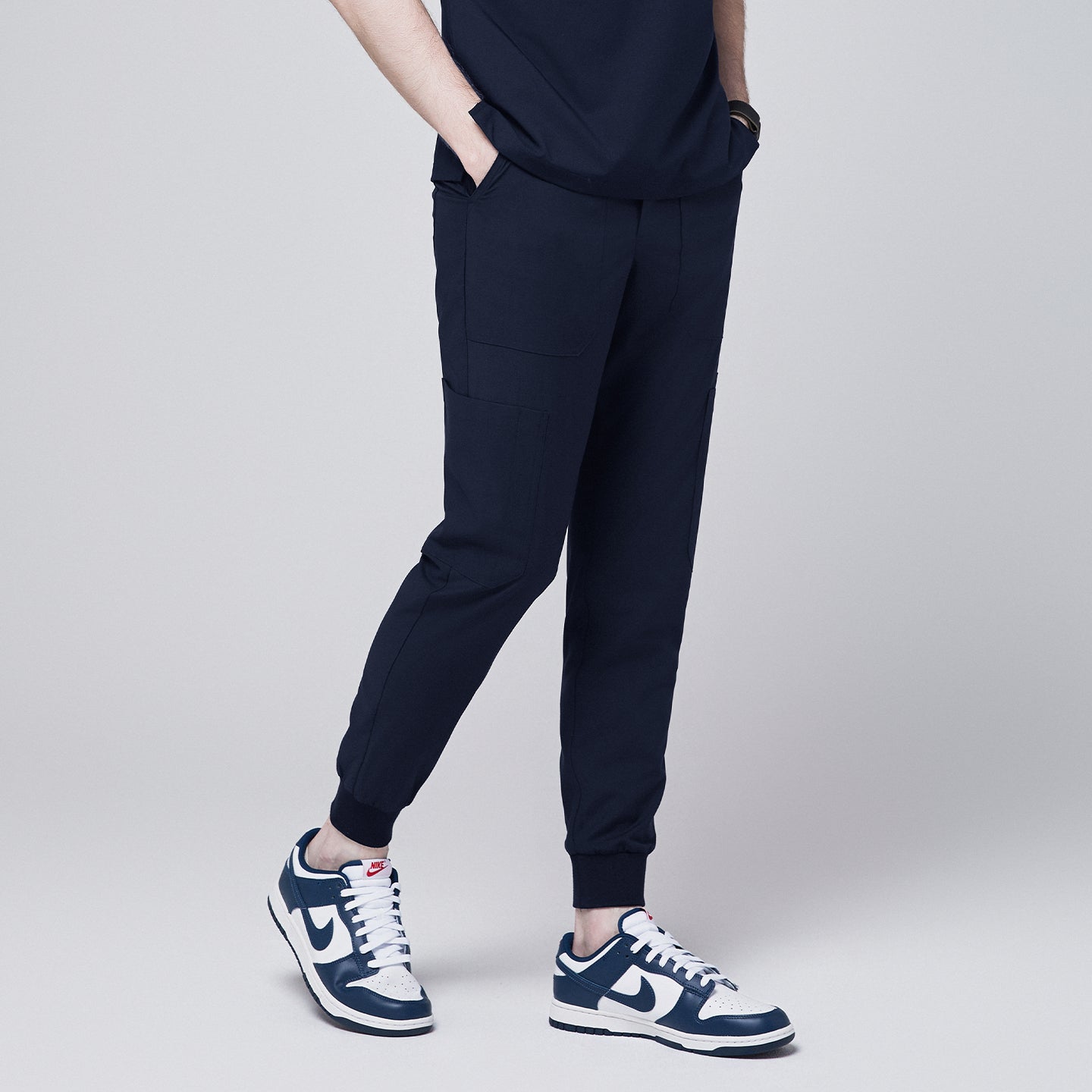 Side angle of navy jogger scrub pants with 8 pockets, drawstring waistband, cuffed ankles, and a name badge, paired with navy sneakers,Eco Navy