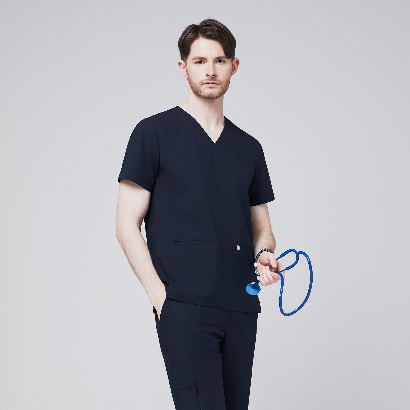 Male model wearing a navy V-neck scrub top with double front pockets, paired with matching scrub pants, holding a blue stethoscope, presenting a professional and modern healthcare uniform style,Eco Navy