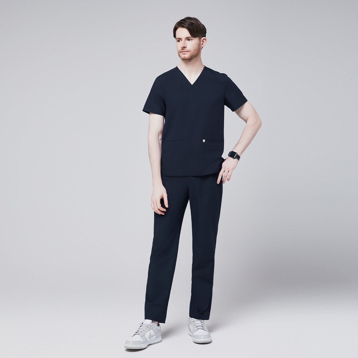 Male model in a full-body pose wearing a navy V-neck scrub top with double front pockets and matching navy scrub pants, styled with casual sneakers, ideal for healthcare professionals,Eco Navy