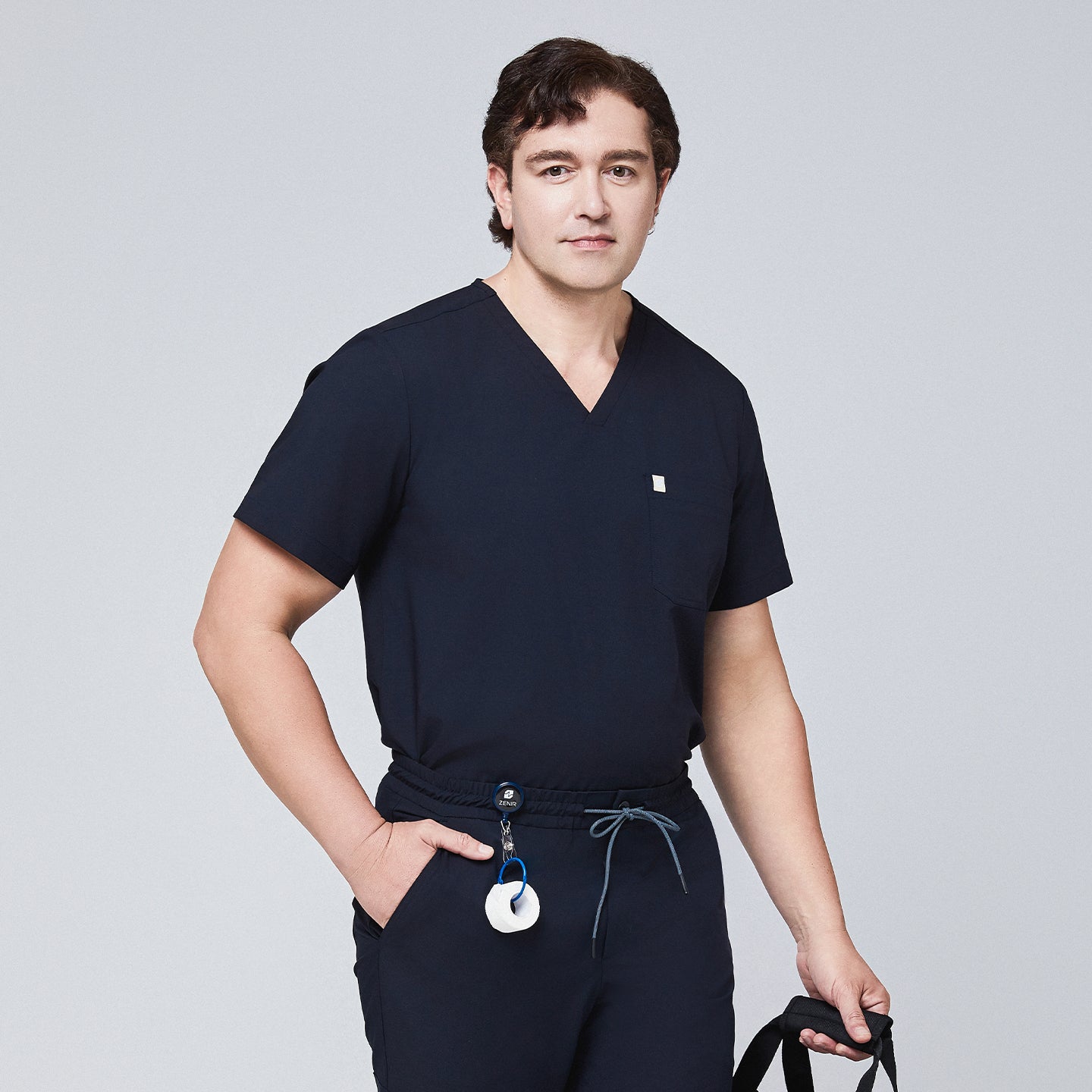 Man wearing a navy scrub set with a single-pocket top, holding a black bag and accessorized with a retractable badge reel,Eco Navy