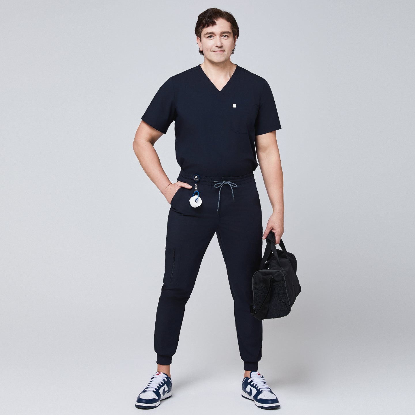 Model wearing navy jogger scrub pants with a zipper pocket and drawstring waist, paired with a matching V-neck scrub top and carrying a black bag,Eco Navy