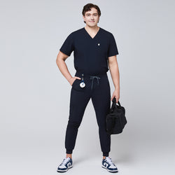 Image of Model wearing navy jogger scrub pants with a zipper pocket and drawstring waist, paired with a matching V-neck scrub top and carrying a black bag,Eco Navy