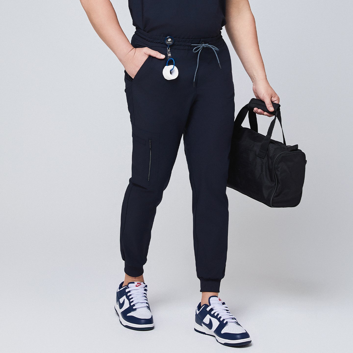 Navy jogger scrub pants with a zipper pocket, drawstring waist, and attached badge clip, styled with a black bag,Eco Navy