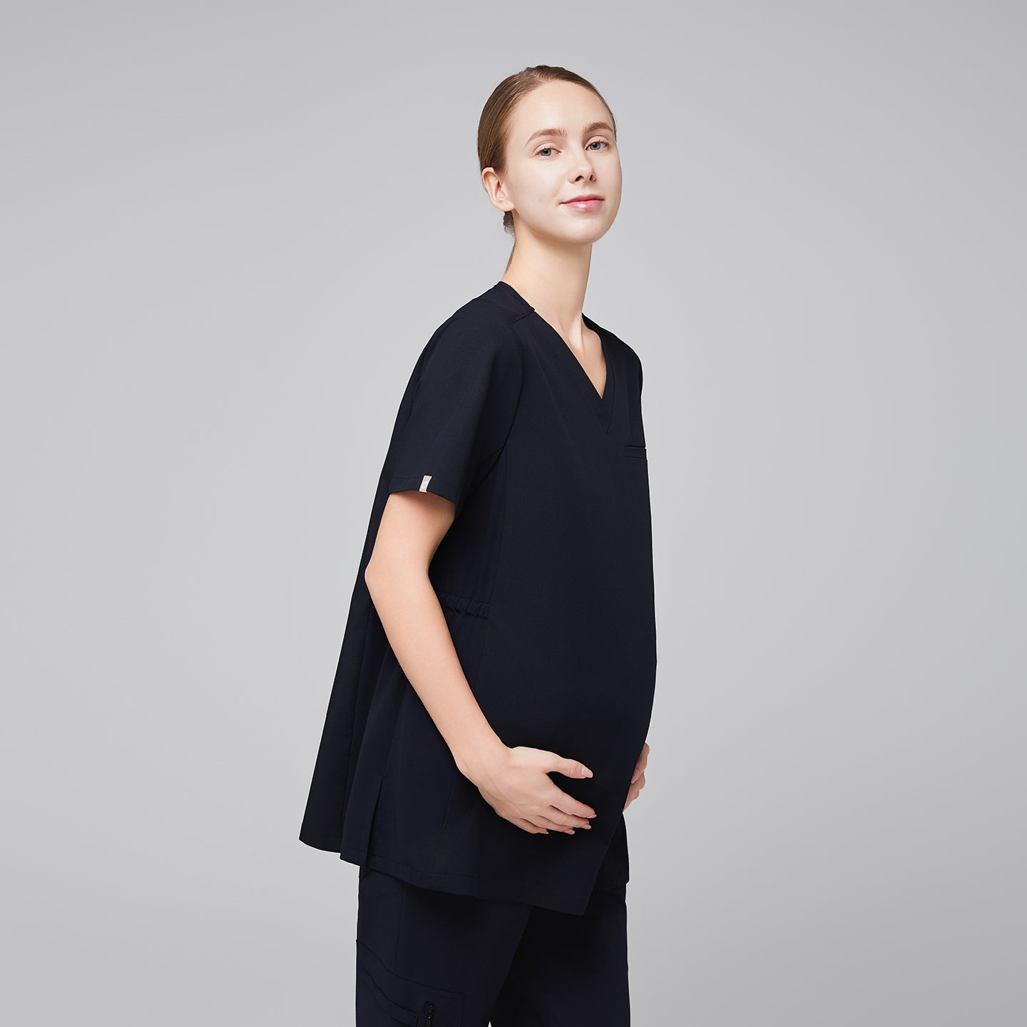 Pregnant woman wearing navy maternity scrub top, shown in a side profile, gently cradling her belly, smiling confidently,Eco Navy