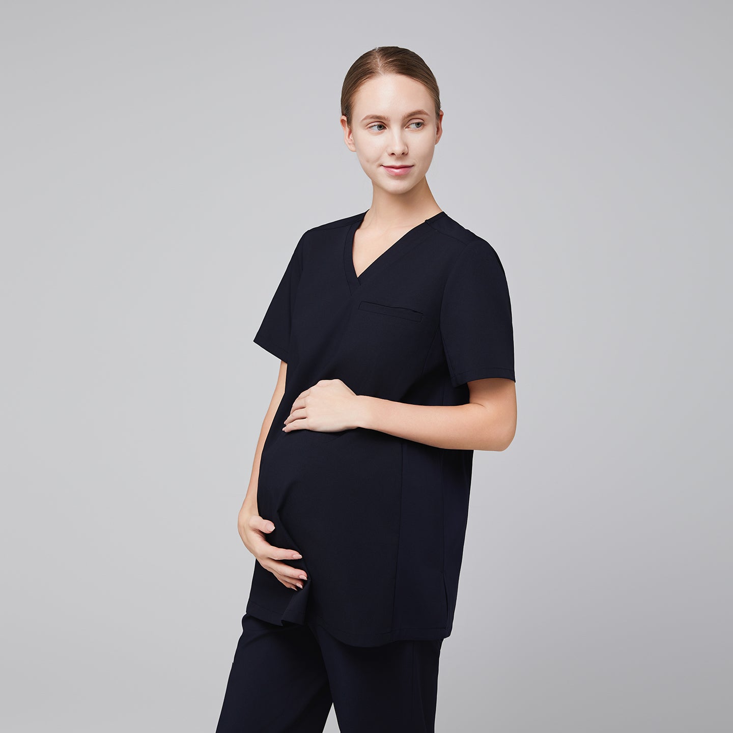 Pregnant woman in navy maternity scrub top, standing in a side pose, gently holding her belly and looking to the side,Eco Navy