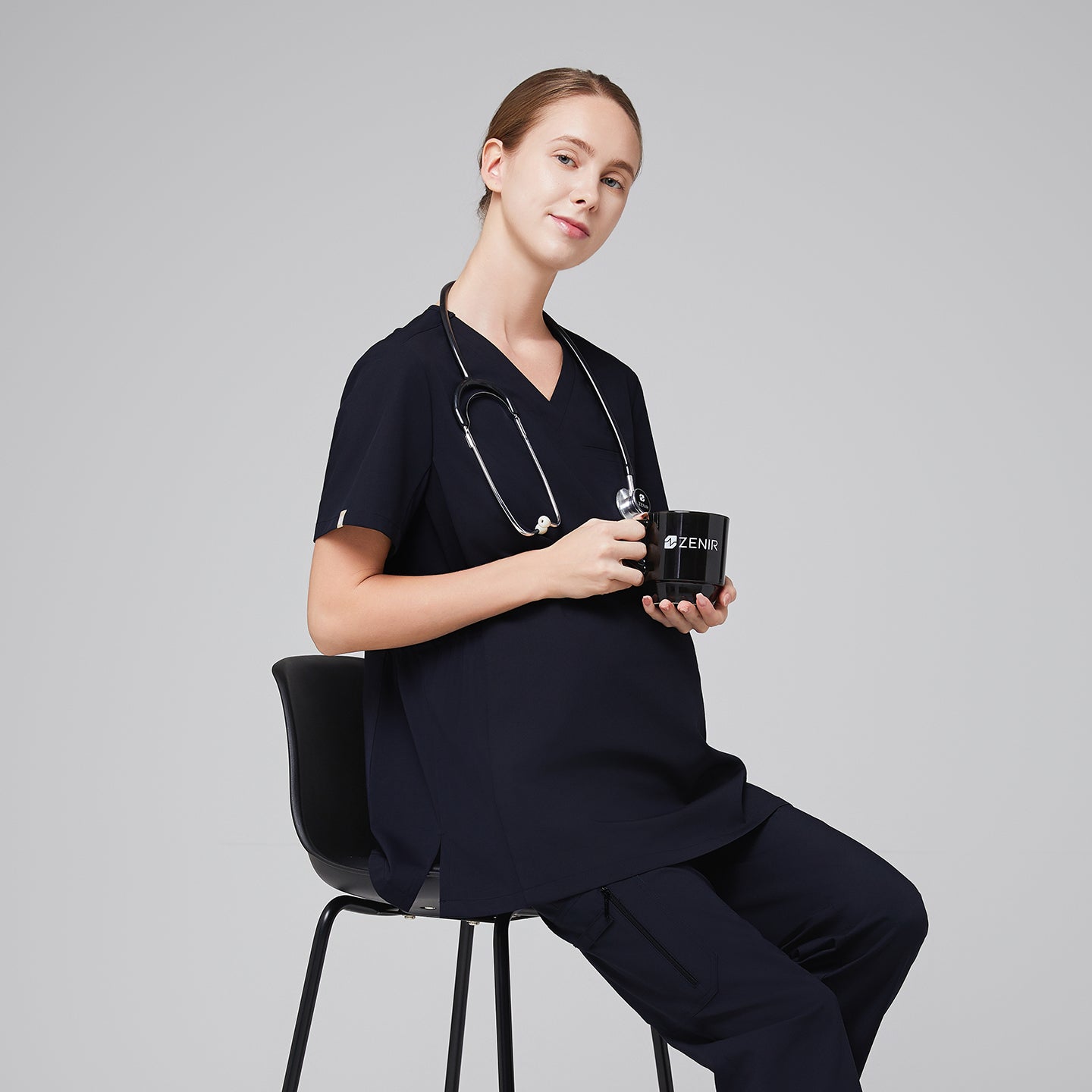 Pregnant woman wearing navy maternity scrub top with a stethoscope around her neck, sitting and holding a ZENIR-branded mug,Eco Navy