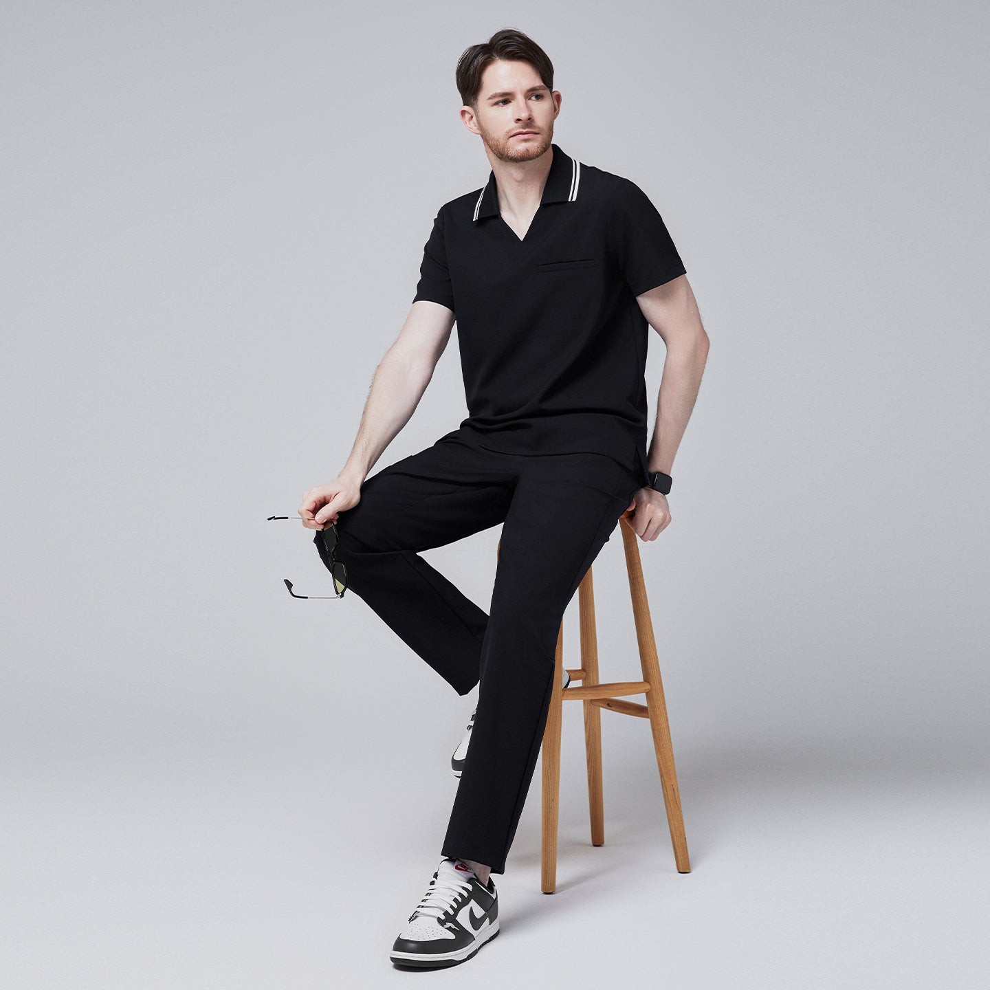 Male model sitting on a wooden stool, wearing a jet black performance polo scrub top with a striped collar, holding sunglasses,Jet Black