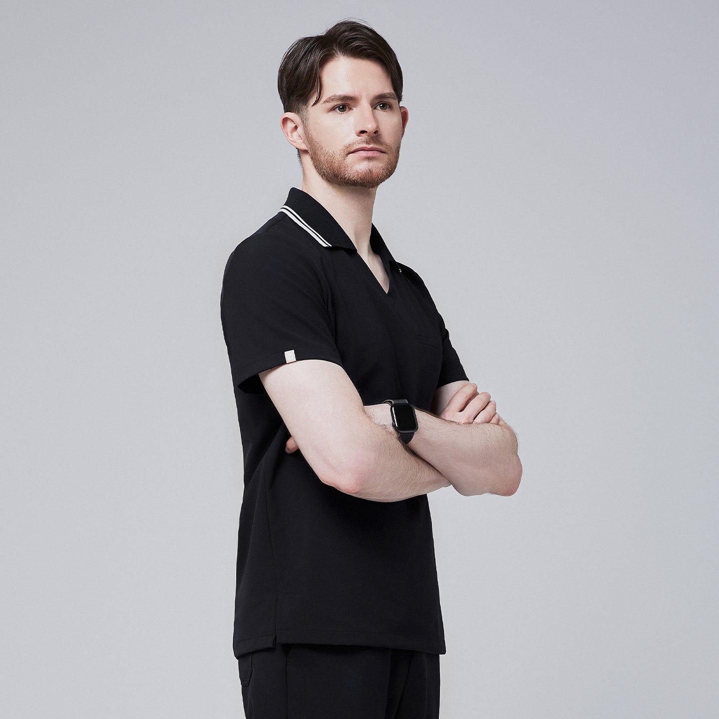 Male model wearing a jet black performance polo scrub top with a striped collar, standing with arms crossed,Jet Black