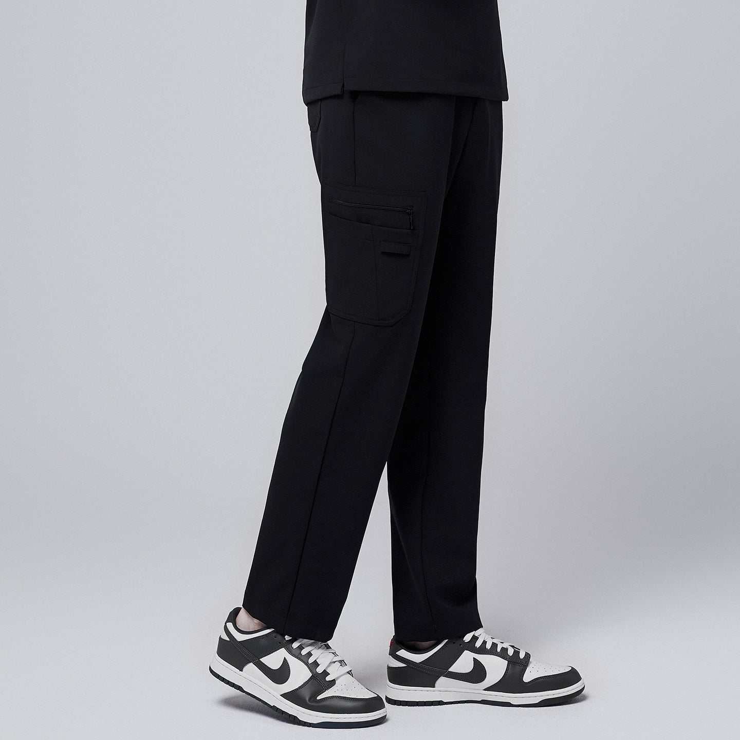 Male model showcasing jet black relaxed-fit scrub pants with functional side pocket design, paired with black and white sneakers, highlighting a professional yet comfortable style,Jet Black